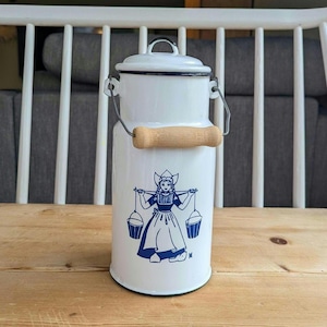 Könnte beinhalten: Weiße Emaille-Milchkanne mit schwarzem Rand und Deckel, mit Holzgriff und einer blau-weißen Illustration einer Frau, die Eimer trägt. Die Kanne hat einen Metallgriff und steht auf einer Holzoberfläche.
