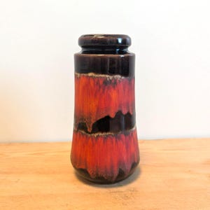 Puede incluir: Un jarrón cilíndrico de cerámica con la parte superior e inferior de color marrón oscuro, que transiciona a un centro rojo y naranja vibrante. El jarrón tiene un acabado brillante y una apariencia texturizada única, ideal para la decoración del hogar.