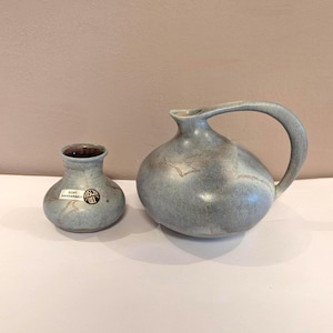 May include: Two ceramic vases in a muted blue-gray hue. One is a small, round bud vase with a dark brown interior and a sticker. The other is a larger pitcher with a curved handle and a rounded body. Both have a speckled appearance.