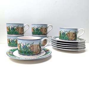 Op de afbeelding: Een set van zes keramische theekopjes en schotels. De kopjes en schotels hebben een lichtblauwe achtergrond met een junglescène, inclusief een olifant, en kleurrijke bloemen- en plantendetails. De kopjes hebben witte handvatten en randen.