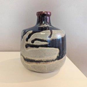 Könnte beinhalten: Eine Keramikvase mit rundem Körper und schmalem Hals. Die Vase hat eine dunkelblaue Glasur mit cremefarbenem Muster und einem rotbraunen Rand. Das Design ist abstrakt und organisch und verleiht dem Stück eine einzigartige Note.