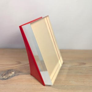 Peut inclure: Un cadre photo en acrylique rouge et transparent avec un intérieur beige clair. Le cadre est incliné et repose sur une surface en bois. Le cadre a un design moderne et minimaliste.