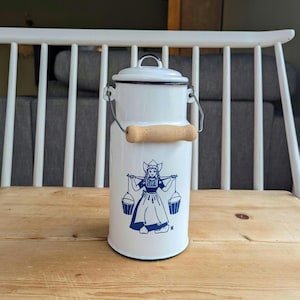 Może przedstawiać: Biały emaliowany dzbanek na mleko z niebieskim wzorem holenderskiej dziewczyny. Dzbanek ma drewniany uchwyt i metalowy uchwyt druciany. Pokrywa jest biała, a dzbanek cylindryczny. Około 25 cm wysokości.