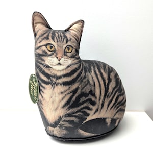 Könnte beinhalten: Ein kissenförmiges Kissen in Form einer Katze mit einem realistischen Tabby-Katzen-Design. Das Kissen hat ein beige-schwarzes Streifenmuster mit gelben Augen und einem detaillierten Gesicht. Ein grünes Etikett ist an der Seite befestigt. Das Kissen ist so gestaltet, dass es wie eine sitzende Katze aussieht.