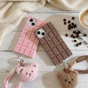Può includere: Due custodie per telefono, una rosa e una marrone, progettate per assomigliare a barrette di cioccolato. Sono presenti anche due portachiavi a forma di orsetto, uno rosa e uno marrone. Una tazza di caffè e chicchi di caffè sono sullo sfondo.