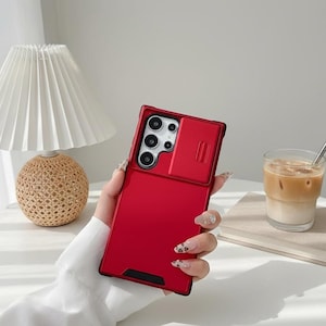 May include: A bright red phone case with a sliding camera cover is held in a hand. The case has a sleek, modern design with a black border. The phone case is shown with a lamp and a glass of iced coffee.