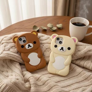 Puede incluir: Dos fundas para teléfono con forma de osos de dibujos animados. Una es marrón con vientre blanco, la otra es color crema con vientre blanco. Ambas tienen ojos negros y nariz negra. Las fundas están sobre una manta de punto color crema.