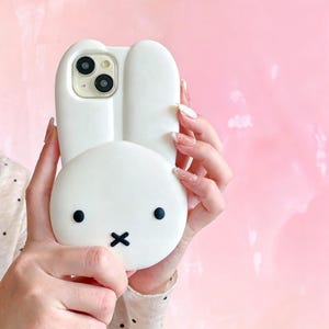 May include: White phone case shaped like a bunny with black eyes and a black 'x' for a mouth. The phone case has a camera cutout and is held against a pink background.