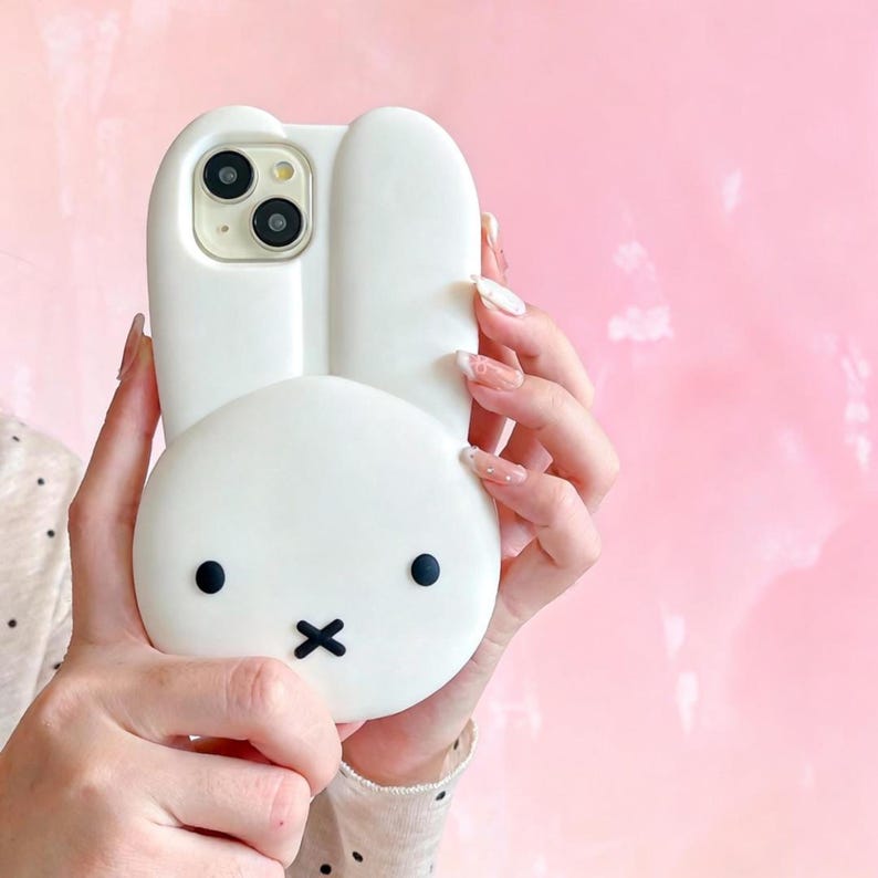 May include: White phone case shaped like a bunny with black eyes and a black 'x' for a mouth. The phone case has a camera cutout and is held against a pink background.
