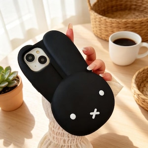 May include: Black phone case with a 3D bunny design. The case features a round head with white dot eyes and a cross for a mouth, and long, upright ears. The phone case is held in a person's hand.