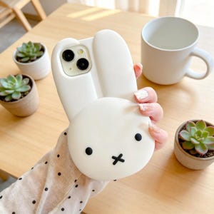 May include: A white phone case featuring a bunny design with black eyes and a cross-shaped mouth. The case has large, upright ears. The phone case is held near small potted succulents and a white mug on a wooden table.