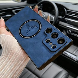 Puede incluir: Funda de teléfono azul con un acabado texturizado y un emblema circular con un diseño estilizado. La funda tiene un borde oscuro y un módulo de cámara con múltiples lentes.