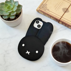 May include: Black phone case with a bunny face design. The case has a rounded shape with white accents for the eyes and mouth. The phone case is on a white marble surface, next to a cup of coffee and a succulent.