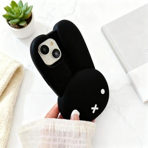 May include: A black silicone phone case shaped like a rabbit head with long ears. The case features two white circular eyes and a white cross-shaped mouth. The camera cutout is positioned at the top, revealing the dual lenses of the smartphone.