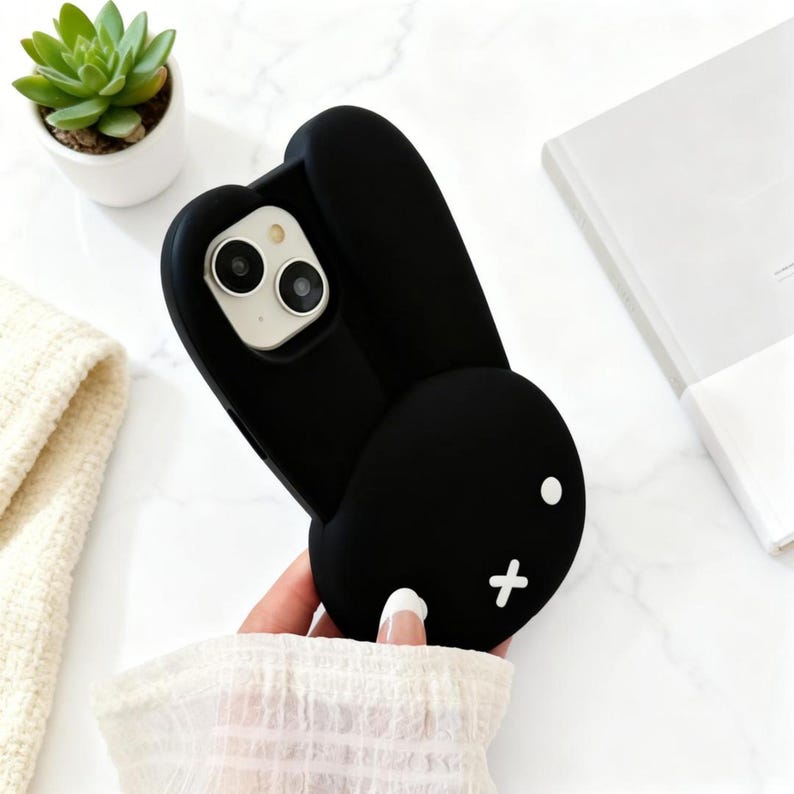 May include: A black silicone phone case shaped like a rabbit head with long ears. The case features two white circular eyes and a white cross-shaped mouth. The camera cutout is positioned at the top, revealing the dual lenses of the smartphone.