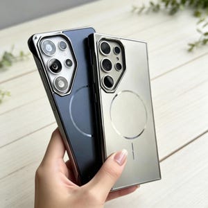 May include: Two phone cases, one navy blue and one silver, are displayed. Both cases have a metallic frame and a circular design on the back. The camera cutouts are visible, and the cases appear to be made of a durable material.