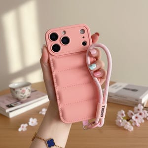 May include: A pink phone case with a quilted design and a matching pink lanyard. The case has a camera cutout and the word "TREND" printed on the lanyard. The case is designed to protect the phone.