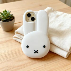 May include: A white smartphone case shaped like a rabbit head with two long ears extending from the top. The case features a minimalist face design with two small black circular eyes and a black cross-shaped mouth centered on the lower portion.