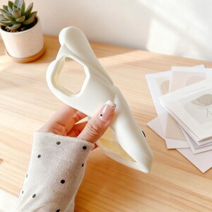 May include: A white phone case in the shape of a whale, held in a hand. The case has a cutout for the camera and buttons. The whale design features a smooth, curved body and a detailed mouth.