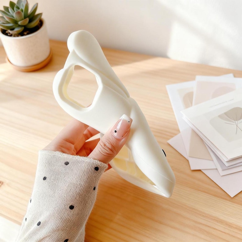 May include: A white phone case in the shape of a whale, held in a hand. The case has a cutout for the camera and buttons. The whale design features a smooth, curved body and a detailed mouth.