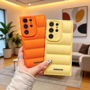May include: Two Samsung phone cases in bright orange and yellow. The cases have a quilted design and a camera cutout. The orange case is held in a hand with a silver ring. The yellow case is held in a hand with a manicure. Both cases have the Samsung logo.