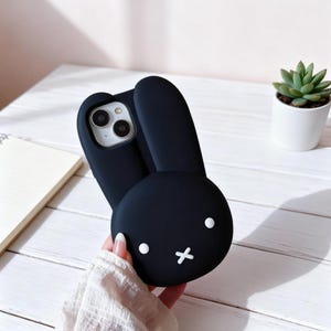 May include: Black phone case with a bunny design. The case features a bunny face with white dot eyes and a white cross for a mouth. The bunny ears extend upwards. The phone case is held in a person's hand.