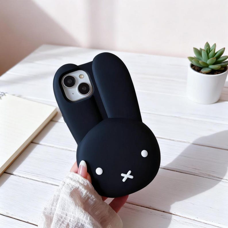 May include: Black phone case with a bunny design. The case features a bunny face with white dot eyes and a white cross for a mouth. The bunny ears extend upwards. The phone case is held in a person's hand.