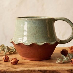 Könnte beinhalten: Eine handgefertigte Keramik-Tasse mit einem einzigartigen Design. Die Tasse hat ein zweifarbiges Farbschema, mit einer helltürkisen oberen Hälfte und einer rotbraunen unteren Hälfte. Der Rand hat einen wellenförmigen, gewellten Rand. Die Tasse steht auf einer Holzoberfläche mit dekorativen Elementen.