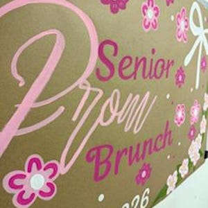 May include: A brown sign with the words "Senior Prom Brunch 2026" in pink and white lettering. The sign is decorated with pink and white flowers and a white bow.