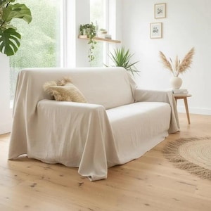 Op de afbeelding: Een beige bank bedekt met een bijpassende linnen hoes. Een getextureerd kussen ligt op de bank. De kamer heeft houten vloeren, een rond jute vloerkleed en een kleine bijzettafel met een vaas met droogbloemen.