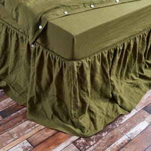 May include: An olive green bed skirt with a ruffled design. The bed skirt is made of a soft, textured fabric and is draped over the bed frame. The bed is covered with a matching olive green sheet and duvet cover. The bed is on a wooden floor.