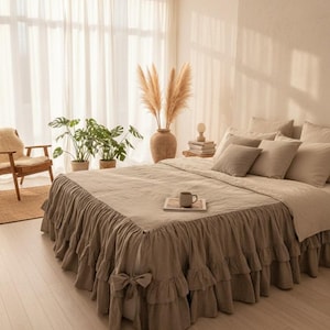 May include: A bed with a neutral-toned linen duvet cover and ruffled bed skirt. The bed is adorned with several matching pillows. A small tray with a mug sits on the bed. The room is decorated with plants, a vase of dried flowers, and a wooden chair.