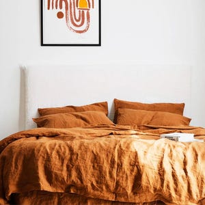 Può includere: Un letto con biancheria da letto in lino color ruggine, tra cui un copripiumino e federe. Sono visibili anche una testiera bianca e un piccolo comodino in legno con un vaso e una pianta. Una stampa d'arte astratta è appesa al muro sopra il letto.