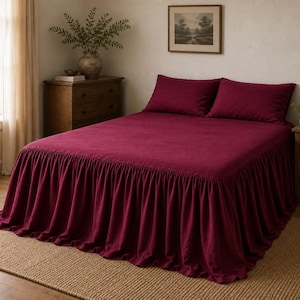 May include: A deep burgundy bedspread with a ruffled skirt that extends to the floor, paired with two matching rectangular pillow shams. The bedding features a solid, matte-textured fabric that covers the entire mattress and frame uniformly.