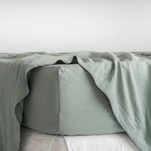 Könnte beinhalten: Ein salbeigrünes Spannbettlaken auf einem Bett. Das Laken besteht aus einem weichen, strukturierten Stoff und hat ein entspanntes, leicht zerknittertes Aussehen. Die Ecke des Lakens ist sichtbar und zeigt das angepasste Design. Darunter ist ein weißes Laken.