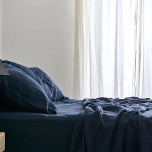 May include: A navy blue linen bedding set, including a fitted sheet, pillowcases, and a top sheet, is arranged on a bed. The sheets have a soft, wrinkled texture. Sheer white curtains are in the background, allowing natural light to filter through.