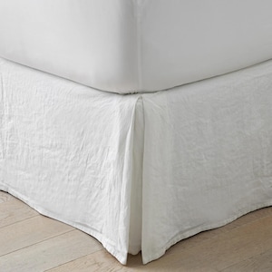 May include: A solid white bed skirt with a textured, slightly wrinkled fabric finish. The bed skirt features a clean, straight-lined design that hangs from the base of the mattress, providing a uniform and neutral appearance for the bed frame.