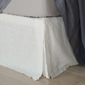 May include: A white bed skirt with a pleated design made of a textured fabric. The bed skirt covers the base of the bed frame, positioned beneath a light gray mattress cover and partially draped by a dark gray fabric hanging from the side.