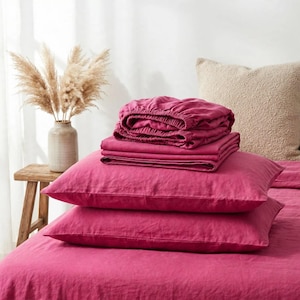 May include: A set of deep magenta bedding including a fitted sheet, a flat sheet, and two pillowcases stacked on a bed. The fabric has a visible woven texture. The bedding is displayed in a bright room with a beige pillow and a vase.