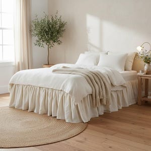 Pode incluir: Uma cama com roupa de cama branca, incluindo uma saia de cama com babados e várias almofadas. Uma manta bege está sobre a cama. Um tapete redondo de juta está no chão de madeira. Uma pequena árvore em vaso é visível.