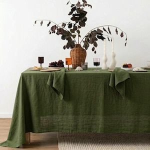 May include: A dining table set for a meal, covered with a green linen tablecloth. The table is decorated with a woven vase holding branches, candles, plates, glasses, and a bowl of grapes. The scene is set against a white wall.