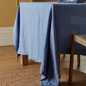 May include: A light blue linen tablecloth draped over a wooden table. The tablecloth has a textured appearance and hangs down, revealing the table legs and a jute rug on the floor. The scene is bathed in natural light.