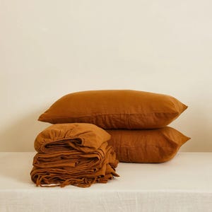 May include: A set of rust-coloured bed linen, including folded sheets and two matching pillowcases. The pillows are stacked. The bedding appears to be made from a soft, textured fabric.