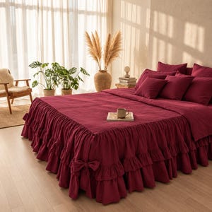 May include: A deep burgundy bedspread featuring a double-layered ruffled skirt with side bow accents. The bedding set includes matching square pillow shams and rectangular pillows, all in a solid, dark red fabric, creating a cohesive look.