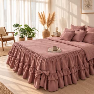 May include: A dusty rose bedspread featuring a double-layered ruffled skirt and a decorative bow detail on the corner. The bedding set includes matching pillow shams in the same muted pink tone, creating a cohesive look for a bed frame.