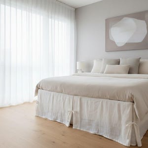 May include: A bed featuring a beige duvet cover and a white bed skirt with tie closures at the corners. The bed is styled with several beige and white pillows. Above the bed hangs a rectangular wall art piece with an abstract white shape.