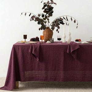 May include: A dining table set with a deep burgundy linen tablecloth. The table is decorated with a woven vase holding dark branches, candles, and glassware. Grapes and small plates complete the elegant setting for a meal.