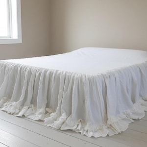 Puede incluir: Una cama blanca con una falda de cama con volantes. La cama está cubierta con una sábana blanca. La habitación tiene pisos de madera pintados de blanco y una ventana en el lado izquierdo. Las paredes están pintadas de un color neutro.