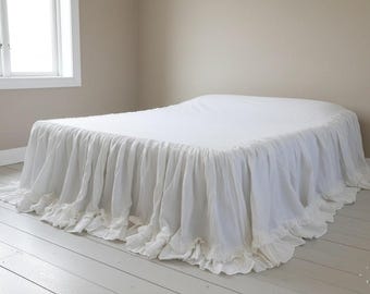 May include: A white bed with a ruffled bed skirt. The bed is covered with a white sheet. The room has white painted wood floors and a window on the left side. The walls are painted a neutral color.