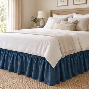 May include: A bed featuring a deep blue ruffled bed skirt that extends to the floor. The bed is dressed in white linens, including a white duvet cover, a light beige throw blanket, and several decorative pillows in shades of white and beige.